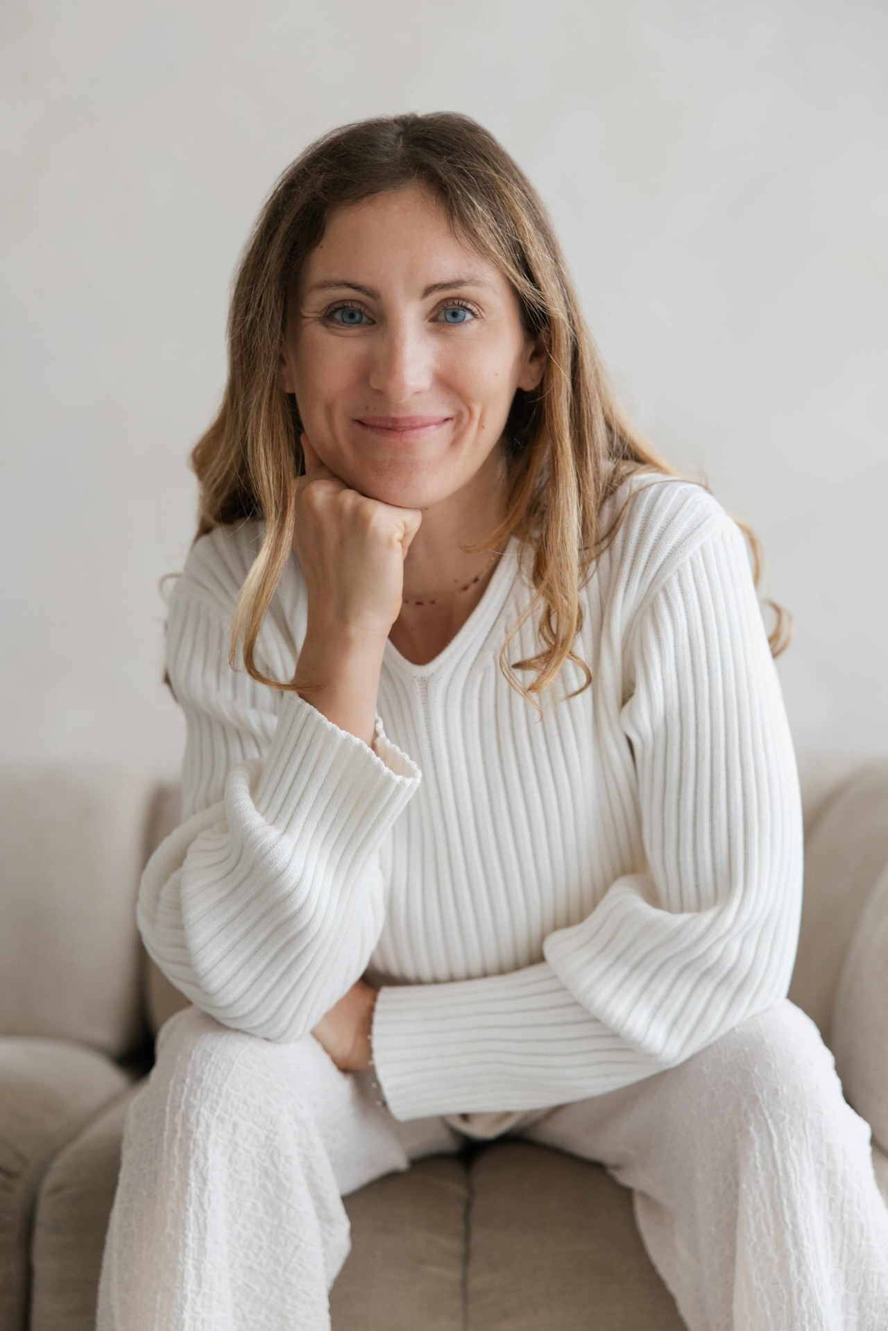 Portrait of Vanessa Michielon seated on a pale sofa in a white ribbed jumper, chin resting on her hand, against a plain light wall